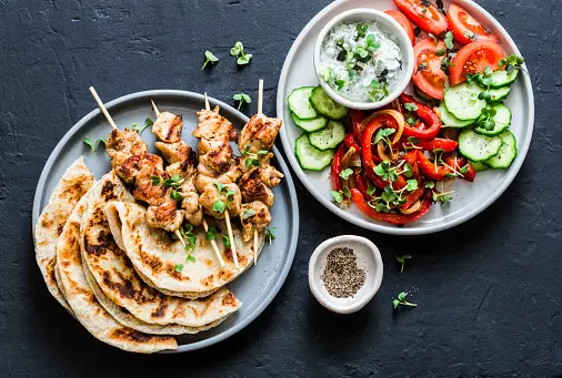 Mediterranean Style Lunch Table Turkey Skewers, Flatbread, Tomatoes, Cucumber Salad, Baked Sweet Pepper, Yogurt Herb Sauce On A Dark Background, Top View